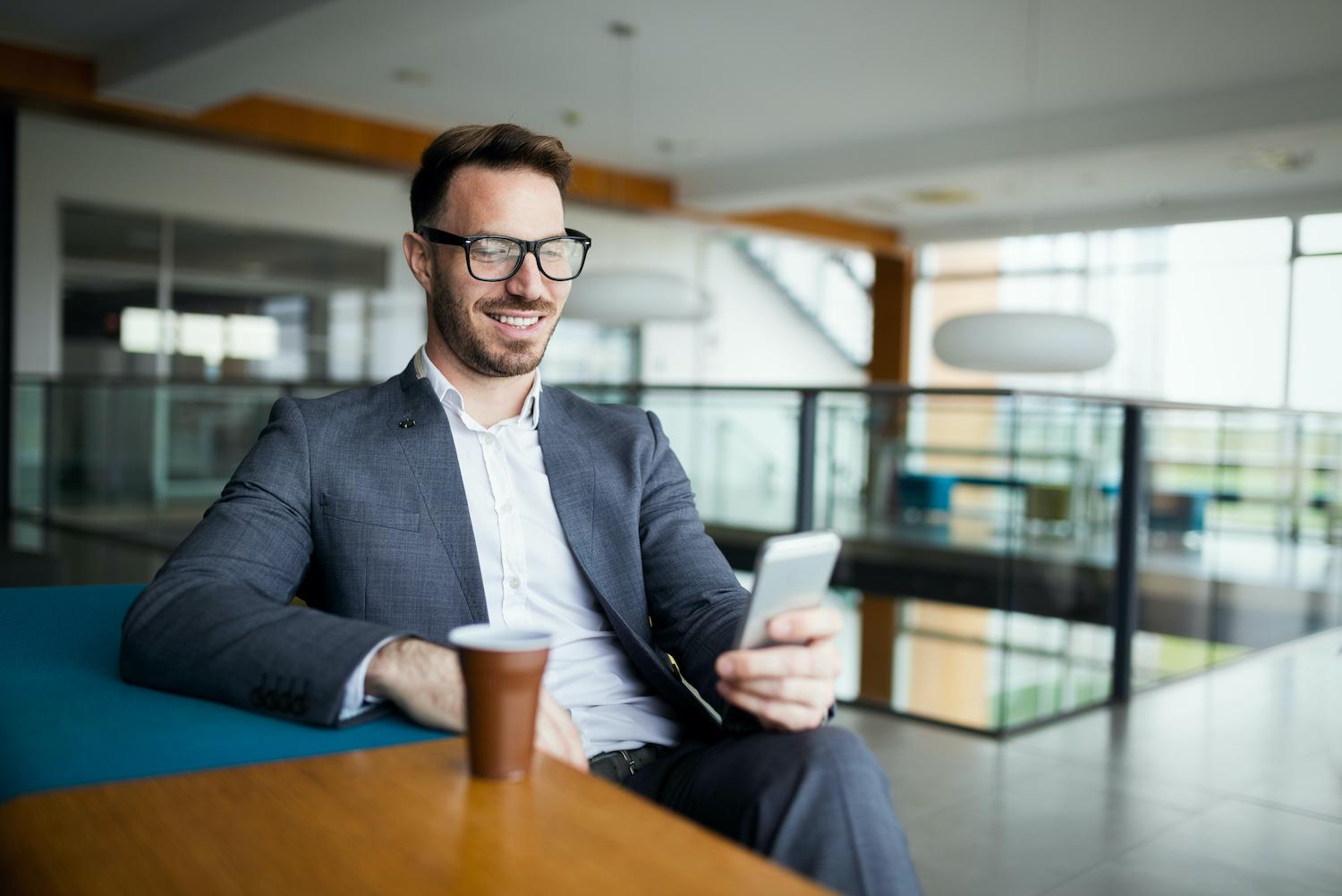A man in a suit sitting a a table with a cup of coffee, looking at his phone and smiling
